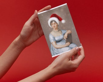 May include: A rectangular greeting card featuring a portrait of a person in a light blue empire-waist dress with a ruffled collar, wearing a red and white Santa hat. The background of the portrait is a soft, muted gray with subtle white snowflakes.