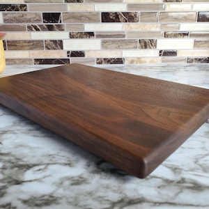 May include: A rectangular, dark brown wooden cutting board with rounded edges. The wood grain is visible, and the board is set on a marble countertop. The board is approximately 18 inches long and 12 inches wide.