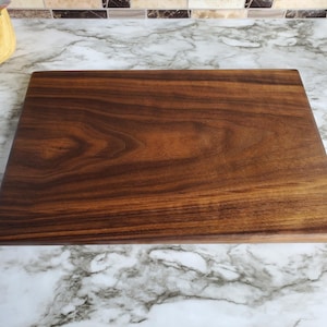 Op de afbeelding: Een rechthoekige houten snijplank met een rijke, donkerbruine kleur en zichtbare houtnerf. De plank is glad en lijkt uit &eacute;&eacute;n stuk hout te bestaan. De snijplank ligt op een marmeren aanrecht.