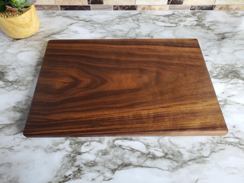 Op de afbeelding: Een rechthoekige houten snijplank met een rijke, donkerbruine kleur en zichtbare houtnerf. De plank is glad en lijkt uit &eacute;&eacute;n stuk hout te bestaan. De snijplank ligt op een marmeren aanrecht.