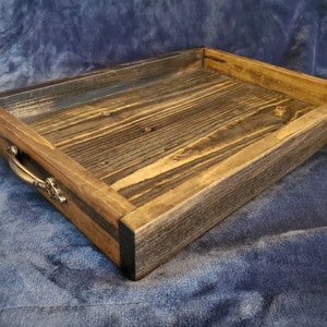 May include: A rustic wooden tray with a dark brown stain and a natural wood grain. The tray has a rectangular shape and features two brass handles on the side.