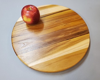 May include: A round wooden cutting board with a natural wood grain pattern. The board is made of various shades of brown wood and has a red apple on top. The cutting board is approximately 12 inches in diameter.