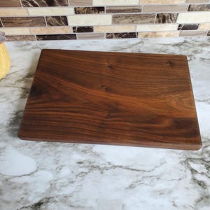 Op de afbeelding: Een rechthoekige, donkerbruine houten snijplank met zichtbare houtnerf. De plank wordt gepresenteerd op een marmeren aanrecht. De snijplank wordt waarschijnlijk gebruikt voor het bereiden en serveren van voedsel.