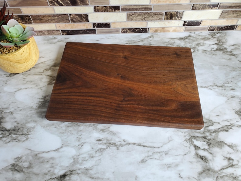Op de afbeelding: Een rechthoekige, donkerbruine houten snijplank met zichtbare houtnerf. De plank wordt gepresenteerd op een marmeren aanrecht. De snijplank wordt waarschijnlijk gebruikt voor het bereiden en serveren van voedsel.