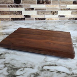 Op de afbeelding: Een rechthoekige, donkerbruine houten snijplank. De plank heeft afgeronde randen en een glad oppervlak, dat de natuurlijke houtnerf laat zien. Ontworpen voor het bereiden en serveren van voedsel, ongeveer 30 cm lang.