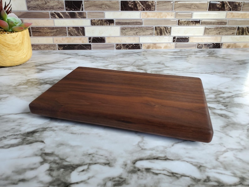 Op de afbeelding: Een rechthoekige, donkerbruine houten snijplank. De plank heeft afgeronde randen en een glad oppervlak, dat de natuurlijke houtnerf laat zien. Ontworpen voor het bereiden en serveren van voedsel, ongeveer 30 cm lang.