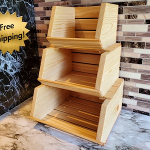 May include: Three-tiered, unfinished wooden storage crates. Each crate is rectangular with a slatted bottom and angled sides. The wood has a light, natural color. The crates are stacked on top of each other.