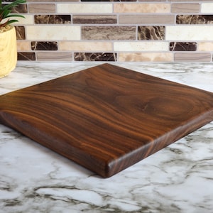 Op de afbeelding: Vierkant houten snijplank, donkerbruin van kleur met zichtbare houtnerf. De plank is gemaakt van massief hout en heeft een glad, afgewerkt oppervlak. De snijplank wordt gepresenteerd op een marmeren aanrecht.