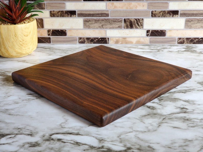 Op de afbeelding: Vierkant houten snijplank, donkerbruin van kleur met zichtbare houtnerf. De plank is gemaakt van massief hout en heeft een glad, afgewerkt oppervlak. De snijplank wordt gepresenteerd op een marmeren aanrecht.