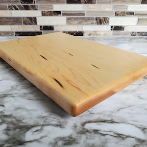 May include: A natural wood cutting board with a light brown color and a smooth, rounded edge. The board has a few small knots and grain patterns.