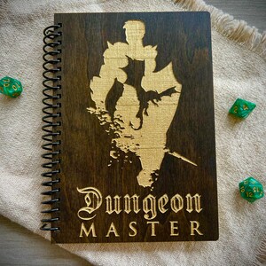 May include: A brown wooden spiral bound notebook with a gold engraved design of a knight and dragon. The notebook has the words "Dungeon Master" engraved on the front.