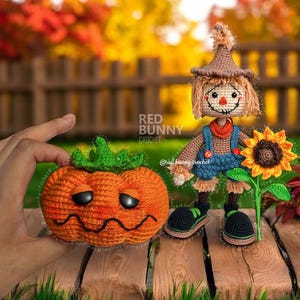 Op de afbeelding: Gehaakte herfstfiguren: een oranje pompoen met groene steel en lachend gezicht, en een vogelverschrikker met bruine hoed en overall, die een zonnebloem vasthoudt. Op de achtergrond een houten hek en herfstbladeren.