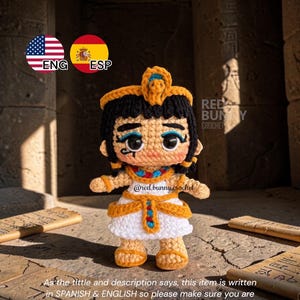 May include: A crocheted doll of an Egyptian figure, featuring a white dress with gold accents, black hair, and a gold headdress. The doll has large, expressive eyes and is standing in front of a stone background. The text "Red Bunny Crochet" is visible.