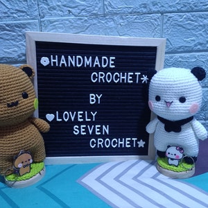 May include: Handmade crochet brown bear and panda plush toys with black eyes and smiling faces. The toys are standing on wooden bases with green grass and smaller versions of the toys. A black letter board behind the toys reads "HANDMADE CROCHET* BY LOVELY SEVEN CROCHET*".