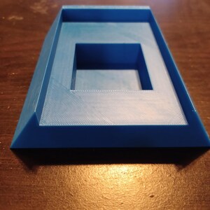 May include: A blue plastic 3D printed stand with a square recess in the center. The stand has a beveled edge and a smooth surface.