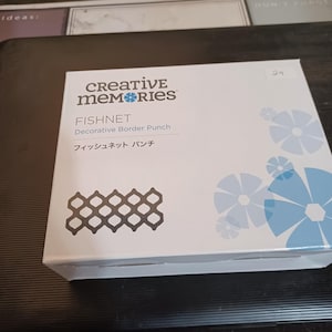 May include: A white box with the text "Creative Memories" in black lettering. The box contains a decorative border punch called "Fishnet". The box also has the text "Decorative Border Punch" and "フィッシュネット パンチ" in black lettering.
