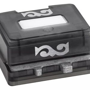 Puede incluir: Un sello rectangular negro con un diseño decorativo plateado en la parte superior y frontal. El sello tiene una ventana rectangular transparente en la parte superior. La base del sello es de metal plateado.