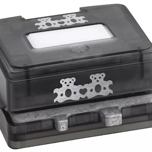 May include: A black paper craft punch with a rectangular shape and a transparent top. The punch features a silver bear design on the front and top. The punch is designed to cut out shapes.