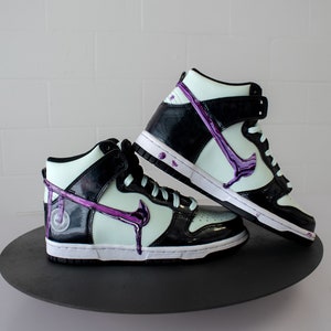 May include: A pair of black and white high-top trainers with a purple swoosh and dripping paint detail. The trainers are on a black circular platform.