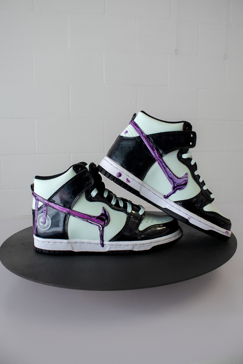May include: A pair of black and white high-top trainers with a purple swoosh and dripping paint detail. The trainers are on a black circular platform.