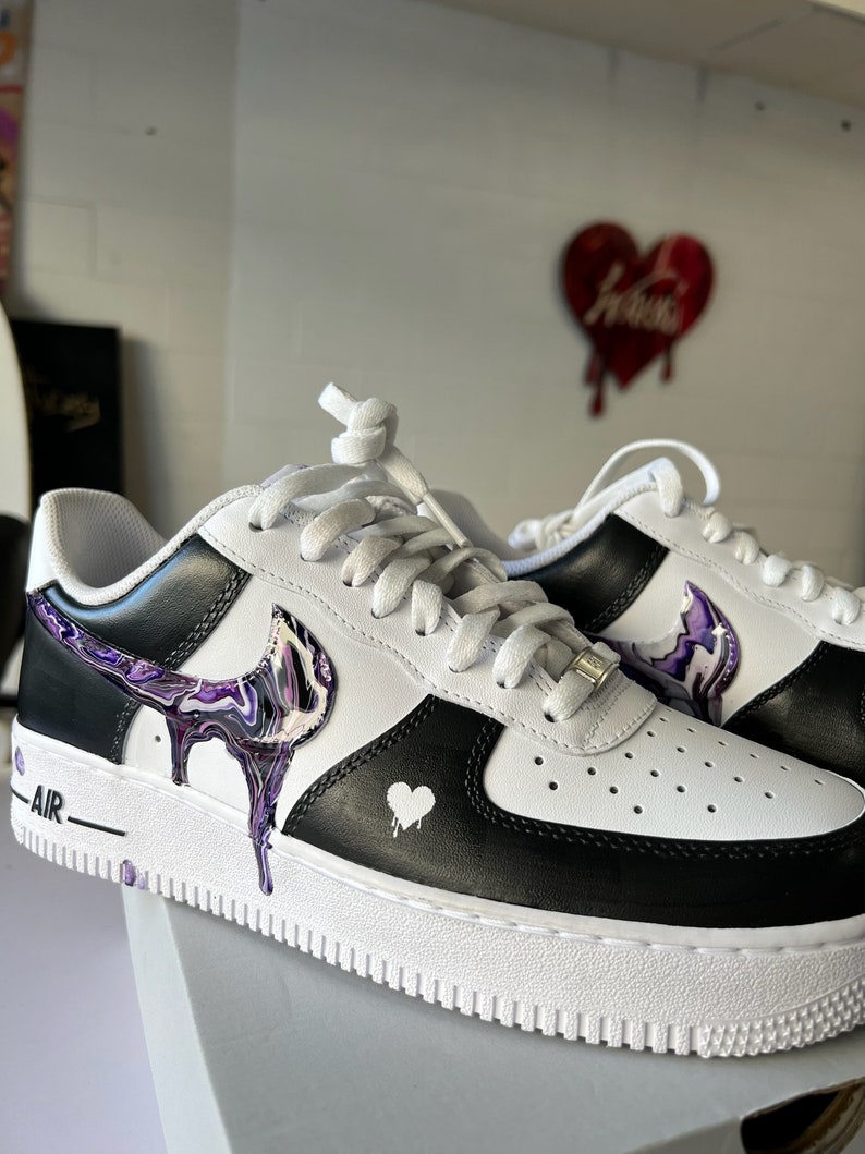 May include: Custom-painted white and black Nike Air Force 1 trainers with a purple and white abstract design on the swoosh and a small white heart on the side. The trainers have white laces and a white sole.