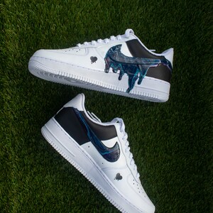 May include: A pair of white trainers with black accents and a blue and black paint design. The paint design resembles a dripping effect.