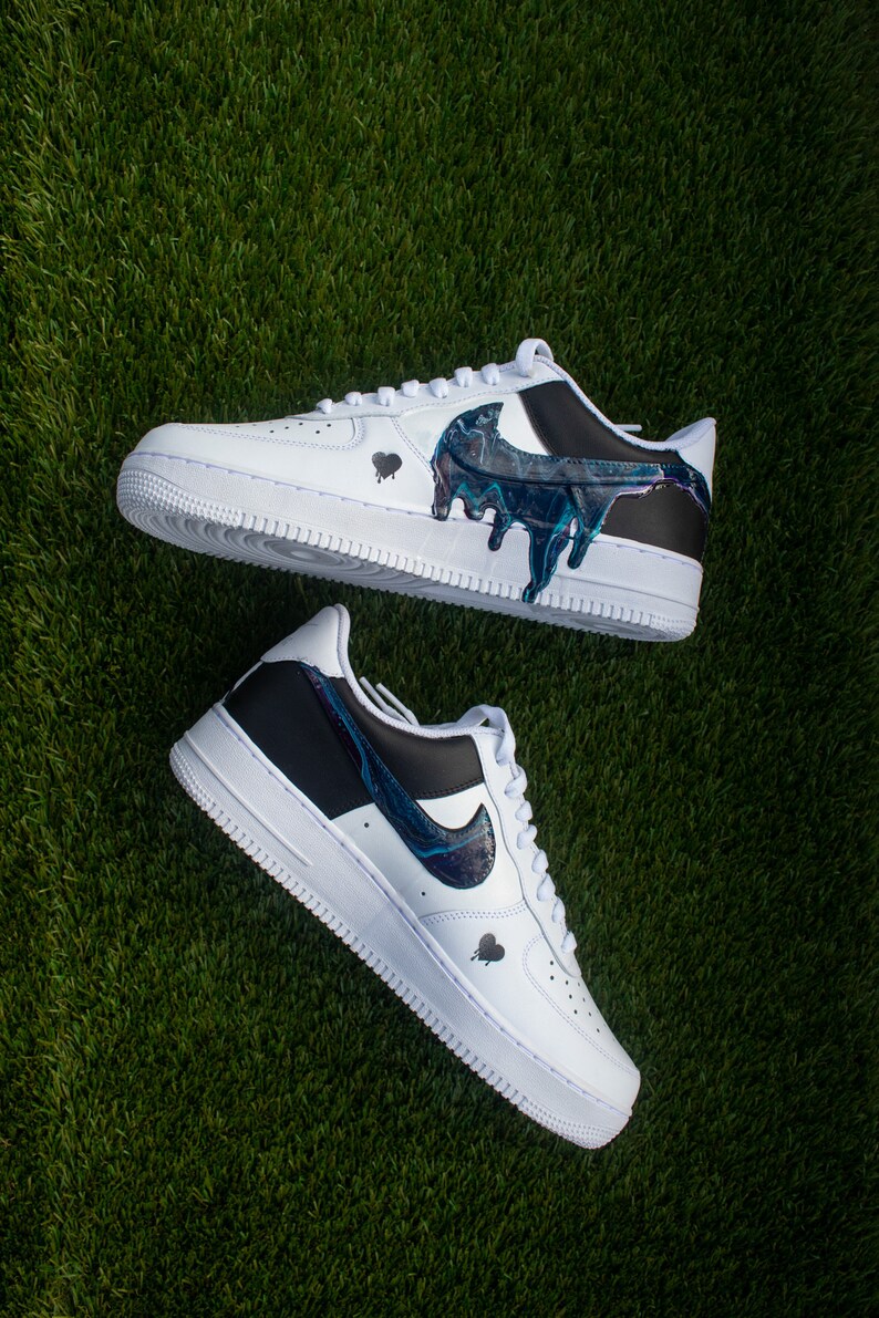 May include: A pair of white trainers with black accents and a blue and black paint design. The paint design resembles a dripping effect.