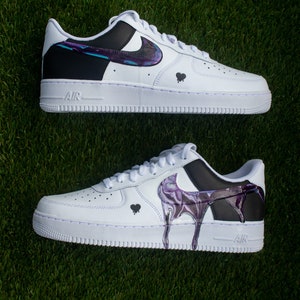 May include: A pair of white Nike Air Force 1 trainers with black and purple paint splatter designs. The trainers have a black swoosh logo and a black heart on each side.