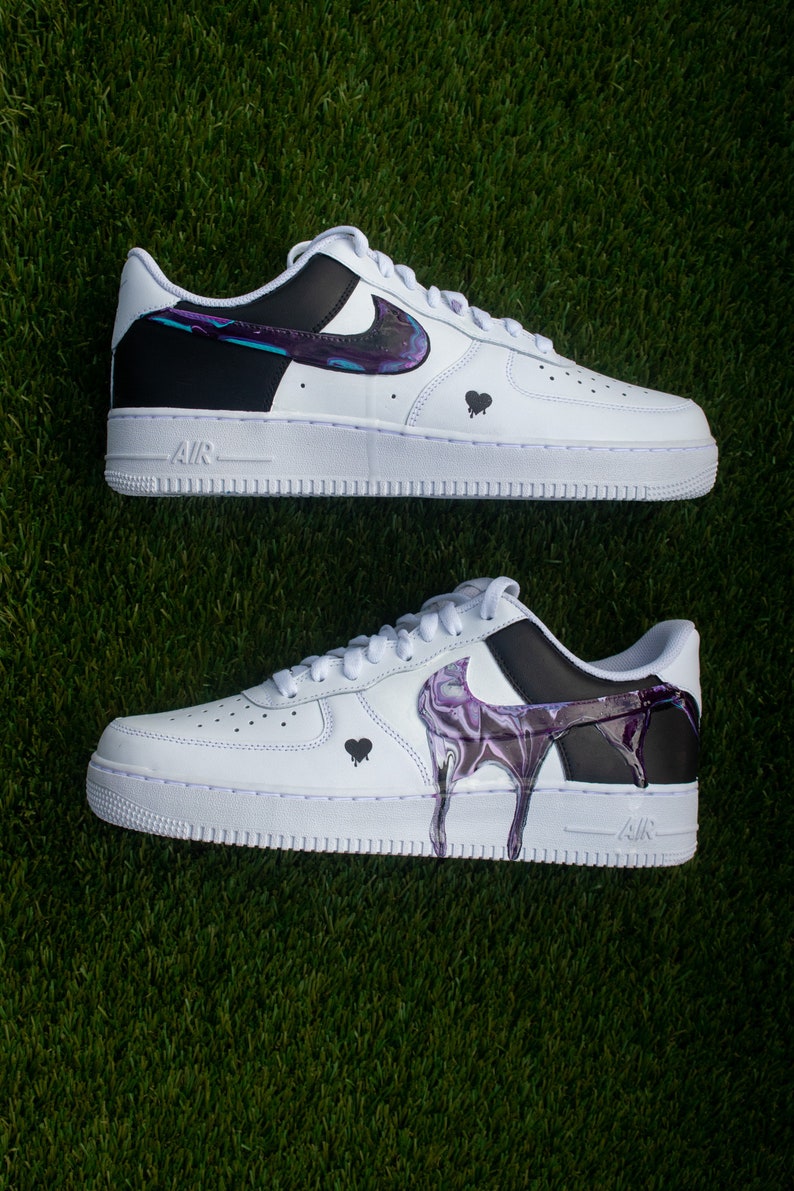 May include: A pair of white Nike Air Force 1 trainers with black and purple paint splatter designs. The trainers have a black swoosh logo and a black heart on each side.