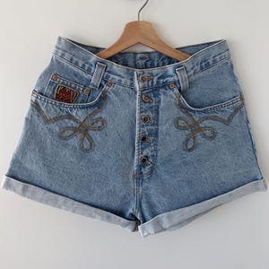 Puede incluir: Pantalones cortos de mezclilla azul claro con cierre de botones y un diseño bordado marrón. Los pantalones cortos tienen un parche en el bolsillo delantero que dice "Wrangler".