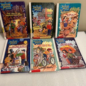 May include: Six children's mystery books from the "Jigsaw Jones Mystery" series. Titles include "The Case of the Haunted Scarecrow," "The Case of the Sneaker Snak," and "The Case of the Bear Scare." Each book features colorful illustrations and the author's name, James Preller.