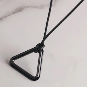 May include: A black metal triangle pendant necklace. The pendant is a hollow, geometric triangle shape, suspended from a thin black cord. The necklace is displayed on a white marble surface.