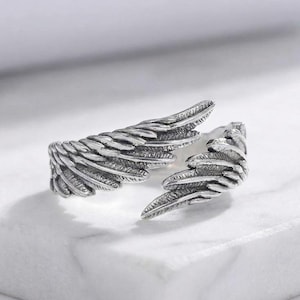 Pode incluir: Um anel prateado em forma de asas de anjo. O anel apresenta texturas detalhadas de penas e um design aberto. As asas envolvem o dedo, criando um acessório único e simbólico.