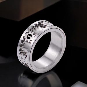 Puede incluir: Un anillo de plata con una banda giratoria que presenta un diseño de engranaje. El anillo está hecho de metal y tiene un acabado liso y pulido.
