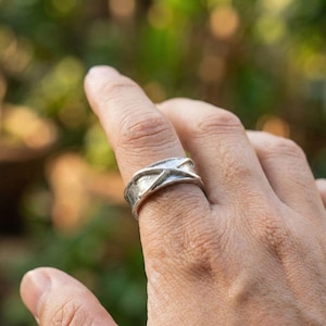Könnte beinhalten: Ein silberner Ring mit einem X-förmigen Design. Der Ring wird an einem Finger getragen, mit einem verschwommenen grünen Hintergrund. Der Ring scheint aus Metall zu sein und hat eine strukturierte Oberfläche.