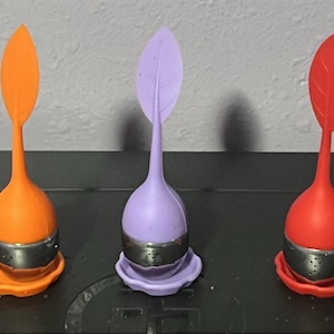 May include: Three colorful tea infusers. Each infuser has a bulbous body, a leaf-shaped top, and a perforated metal basket. The infusers are orange, purple, and red, and sit on matching colored saucers.