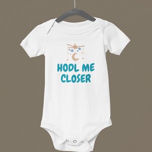May include: White baby onesie with short sleeves, featuring a graphic of a mobile with stars, a moon, and the text "HODL ME CLOSER" in teal. The onesie is hanging on a gray hanger against a brown background.