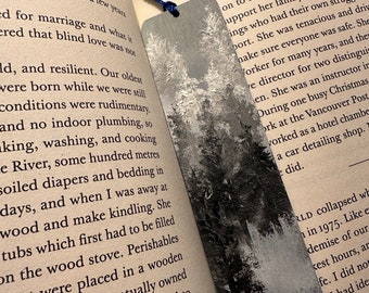 May include: A close-up of a gray and white bookmark with a tree design. The bookmark is made of a textured material and has a blue ribbon attached to it. The bookmark is lying on top of a page of a book with text about a woman's life.