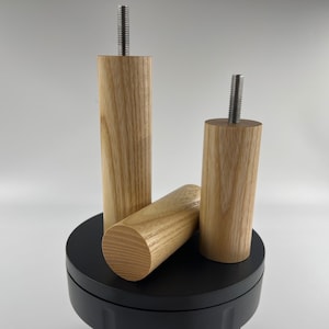 May include: Three unfinished wooden furniture legs with a natural wood grain. Each leg is cylindrical, with a metal threaded bolt protruding from the top. One leg is lying horizontally, while the other two stand upright on a black surface.