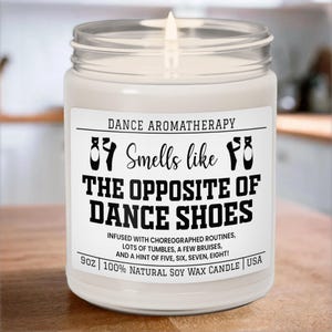 May include: A white soy wax candle with a black label that reads "Dance Aromatherapy 87 Smells like 78 The Opposite of Dance Shoes" and "Infused with choreographed routines, lots of tumbles, a few bruises, and a hint of five, six, seven, eight! 9oz 100% Natural Soy Wax Candle | USA".