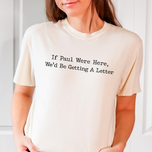May include: Cream-colored t-shirt with the text "If Paul Were Here, We'd Be Getting A Letter" in black. The shirt is made of a soft material and is being worn.