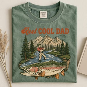 May include: Green t-shirt with the text "Reel Cool Dad" in orange. The graphic features a fisherman in a river, mountains, and a large fish. The shirt is a Comfort Colors brand.