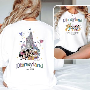 Puede incluir: Sudadera blanca con un gr&aacute;fico de Disneyland colorido en la espalda. El gr&aacute;fico presenta a Mickey Mouse, Minnie Mouse, Donald Duck, Goofy y Pluto frente al Castillo de la Bella Durmiente. La parte delantera de la sudadera tiene un gr&aacute;fico de Disneyland m&aacute;s peque&ntilde;o con el texto "Disneyland est. 1955".