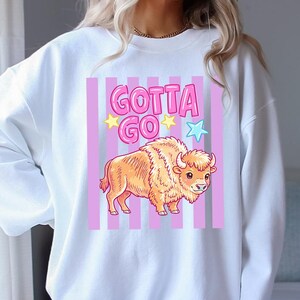 May include: White sweatshirt featuring a cartoon bison with the text "GOTTA GO". The bison is orange and brown, set against a pink and white striped background. The text is in pink with yellow and blue star accents.