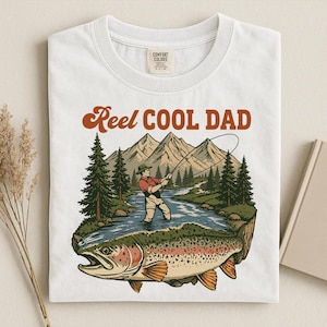 May include: White t-shirt with a graphic of a fisherman in a river, mountains, and a large fish. The text "Reel Cool Dad" is in brown script above the graphic. The shirt has a Comfort Colors tag.