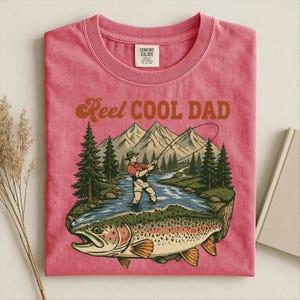 May include: Pink t-shirt with a graphic of a fisherman in a river, mountains, and a large trout. Text reads "Reel Cool Dad" in brown lettering. The shirt has a crew neck and a Comfort Colors tag.