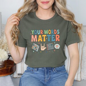 May include: Olive green t-shirt with the text "YOUR WORDS MATTER" in colorful retro font. The shirt features graphics of a calculator, a hand signing "I love you", a keyboard, and a heart speech bubble.