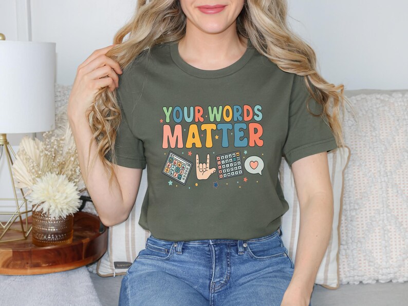 May include: Olive green t-shirt with the text "YOUR WORDS MATTER" in colorful retro font. The shirt features graphics of a calculator, a hand signing "I love you", a keyboard, and a heart speech bubble.