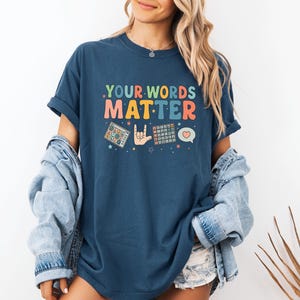 May include: A navy blue t-shirt with the text ".YOUR WORDS MATTER" in colorful retro font. The shirt features graphics of a puzzle, a hand making a rock on gesture, a calculator, and a heart-shaped speech bubble.