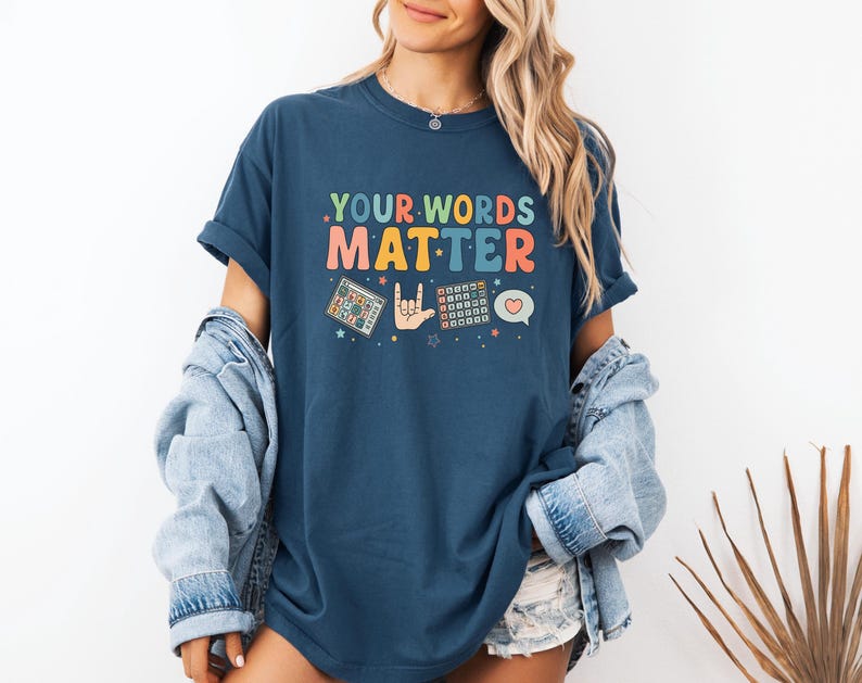 May include: A navy blue t-shirt with the text ".YOUR WORDS MATTER" in colorful retro font. The shirt features graphics of a puzzle, a hand making a rock on gesture, a calculator, and a heart-shaped speech bubble.
